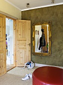 A corner of a room with a red pouffe and a gold framed mirror on a grey wall and an open door