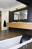 Designerbad mit im Boden eingelassener Badewanne, Waschtisch aus Holz, Badspiegel vor schwarzer Wand und mit indirekter Beleuchtung