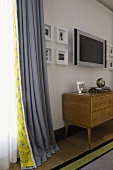 1950's wooden chest of drawers and flat screen TV in front of a wall next to gray curtains