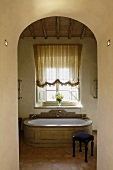 Blick durch Rundbogen auf antike Badewanne in eleganter Steinausführung und Hocker mit blauem Polster