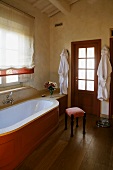 Country style bathroom -- bathtub clad in wood in front of a window with a Roman blind and upholstered stool on a wooden floor