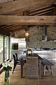 Renovated country home - dining area under a rustic wooden beam ceiling and stainless steel kitchen in front of a natural stone wall