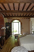 Elegant bedroom in a renovated country home with a rustic wooden beam ceiling and open terrace doors in a round arch