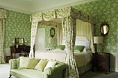 An elegant country house-style bedroom with a green upholstered sofa at the foot of a four-poster bed with a canopy and floral, green wallpapered walls