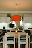A redish orange lampshade hanging over a light wooden dining table and chairs in front of an open-plan, fitted kitchen