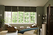 An elegant bedroom with grey wall and a double bed in front a bank of white-painted windows with grey blinds