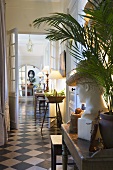 A bust and a palm tree on a wooden wall table in an illuminated corridor of a country house with a black and white tiled floor