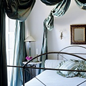 A table lamp with a white shade on a bedside table and an antique metal bed under an elegantly draped canopy
