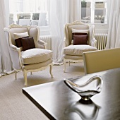 Two light grey, Rococo-style armchairs in front of a window with a light, floor-length curtain