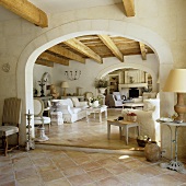 A view through a round archway in a Mediterranean living room with a rustic wooden ceiling, a white sofa and a terracotta floor