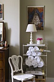 An old chair and a white table lamp and hand towels on metal shelf in front of a grey wall