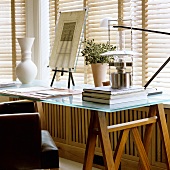 A glass-topped desk with wooden legs in front of a window with a closed blind