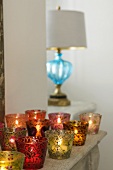 Coloured tea light holders with burning candles on a shelf and a lamp with a blue glass base on the window sill