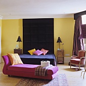 A colourful bedroom - a pink chaise longue with a black bed in the background with a ceiling-high headboard on the yellow wall