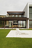 Loungers on a white stone patio in a garden of a house with lots of windows and a wooden porch