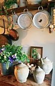 Antique metal colanders hanging from a shelf and handmade clay pots on a wooden shelf below