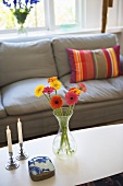 Blumen in Vase und Kerzenständer auf Couchtisch vor grauem Polstersofa mit gestreiftem Kissen