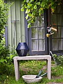 Blaue Laterne auf Steinbank und Schale mit Gartengeräten vor Gartenhäuschen