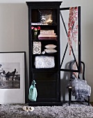 Black ceiling high display case with open door with odds and ends inside next to an antique chair in front of a gray wall