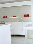 Modern white kitchen with red glassware on shelf above cupboard units.