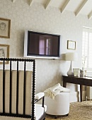 A detail of a traditional study room decorated in neutral colours, pattern wallpaper, beamed ceiling, wooden chair with cushions, ottoman, wall mounted television, table, lamp,