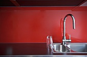 Glas auf Spülbecken mit Armatur vor rotem Spritzschutz