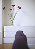 A detail of a modern, white bedroom, upholstered headboard in foreground, arrangement of Allium flowers in glass vase on unit,