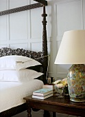 A detail of a traditional bedroom with painted panelling, carved wooden four poster bed, bedside table, lamp