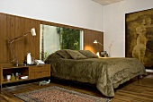 A double bed with a green fur cover in front of a window and a bedside table in front of a wooden wall in the same wood