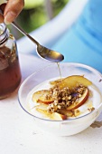 Young woman pouring honey over yoghurt with nectarines & nuts 