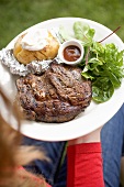 Woman holding plate of grilled beef steak & accompaniments