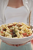 Woman holding a bowl of pasta salad with tomatoes & olives