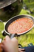 Hand holding a pan of baked beans, barbecue in background