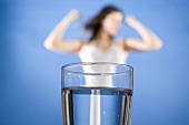 Glass of water, slim woman flexing her muscles in background