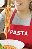 Young woman in apron eating spaghetti with tomato sauce