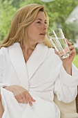 Frau in weißem Bademantel trinkt ein Glas Wasser im Garten