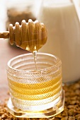 Honig fliesst von Honiglöffel in Honigglas, dahinter Milch