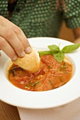 Hand dippt Weißbrot in Tomatensuppe mit Basilikum