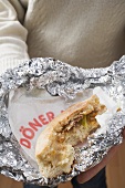 Person holding döner kebab, partly eaten, in aluminium foil