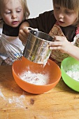 Zwei Kinder bereiten Teig zu (Mehl in Schüssel sieben)
