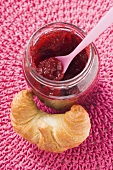 Jar of raspberry jam with spoon, croissant beside it