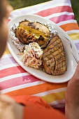 Woman holding plate of grilled steak, baked potato & coleslaw