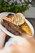 Hands holding plate of steak, bread, corn on the cob, baked beans