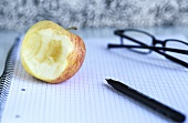 Angebissener Apfel, Brille und Schreibstift auf Notizblock
