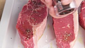 New York strip steaks being seasoned with pepper