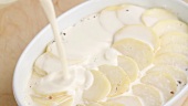 Cream being poured over potato slices in a baking dish (close-up)