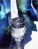 Wasserglas mit perlendem Wasser, im Hintergrund Flaschen