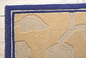 Beiger Wollteppich mit reliefartigem Motiv, blaue Umrandung.
