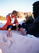 Woman wearing capri jeans and orange Red knee length parka sitting in sand