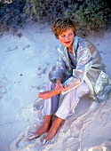 Woman wearing blue pants and transparent nylon parka sitting in sand and looking away